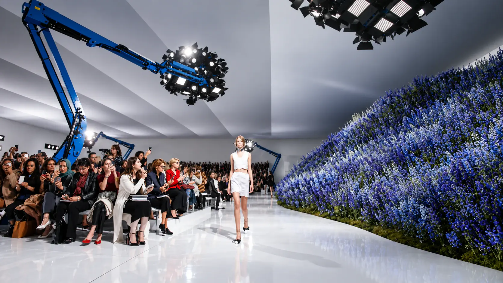 A Dior model walking beside a wall of blue flowers at a runway show