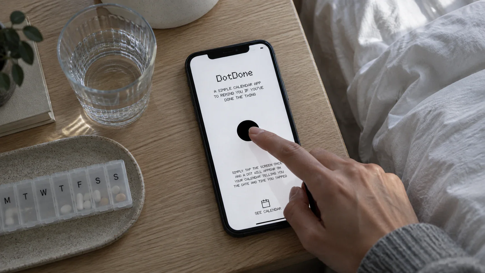 A person recording a DotDone entry beside a glass of water and a weekly pill organiser
